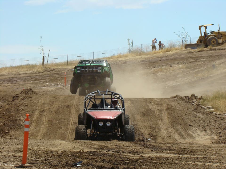 Two off-road buggies racing on a dirt track, kicking up dust. A yellow bulldozer is visible in the background