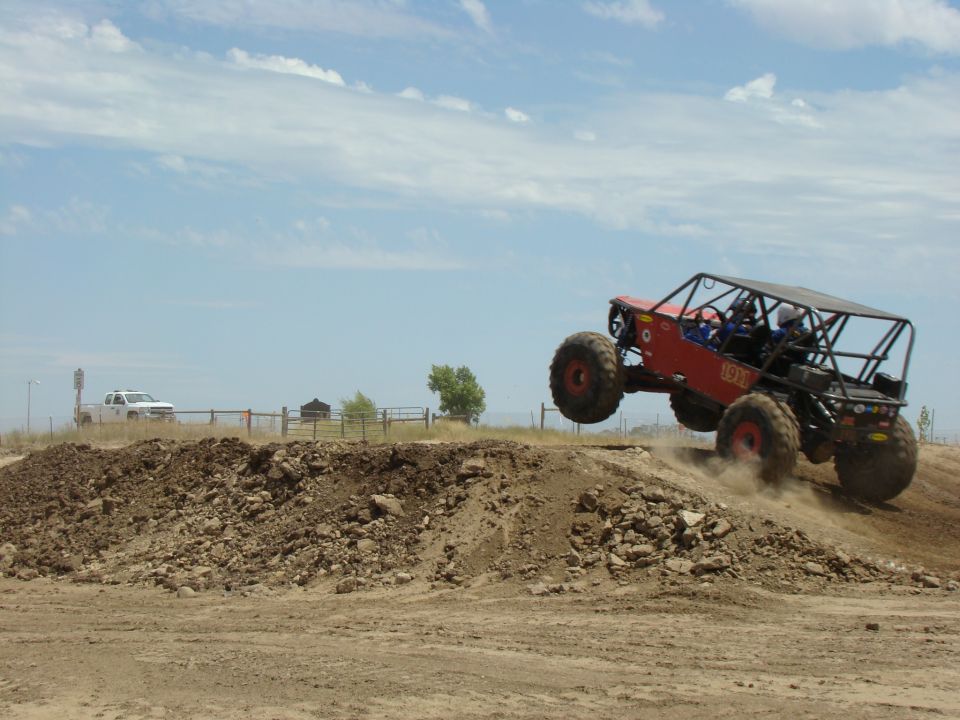 Red off-road buggy with a roll cage is airborne over a dirt mound. A white pickup truck is visible in the background