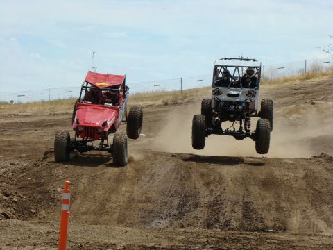 1989 Ford Bronco - Two custom off-road vehicles catch air during a race on a dusty dirt track.