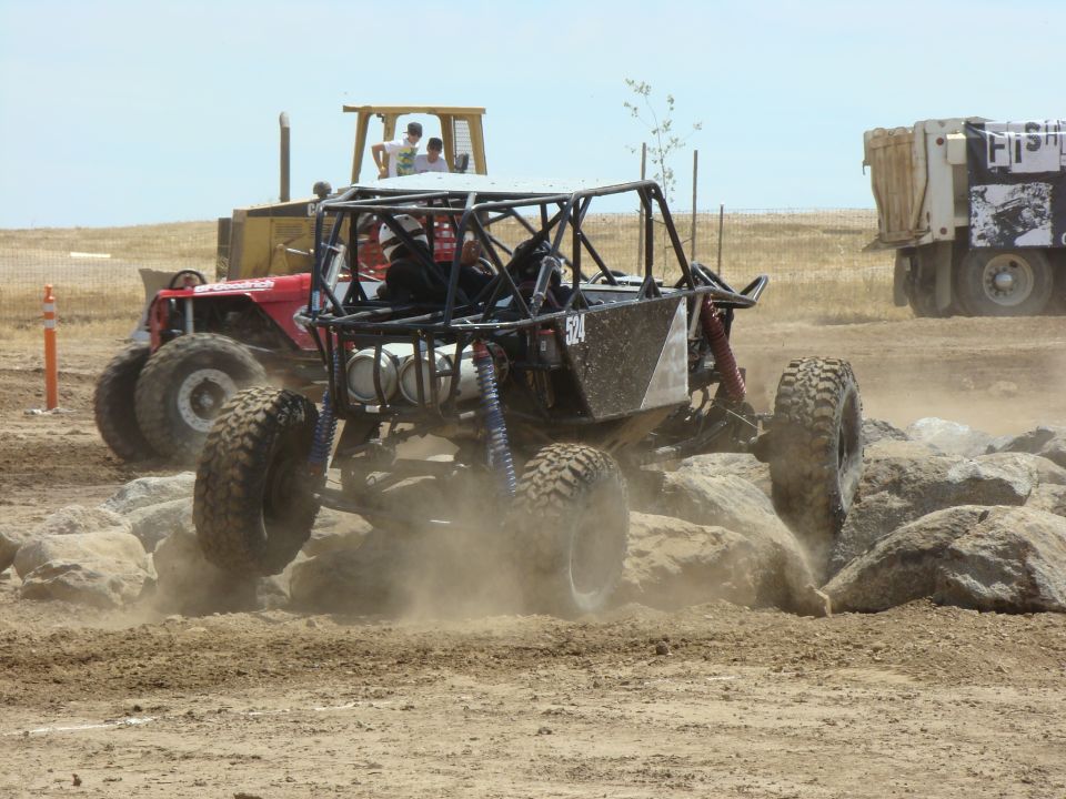 Off-road buggy with number 524 on side, climbing over rocks with a bulldozer and dump truck in background