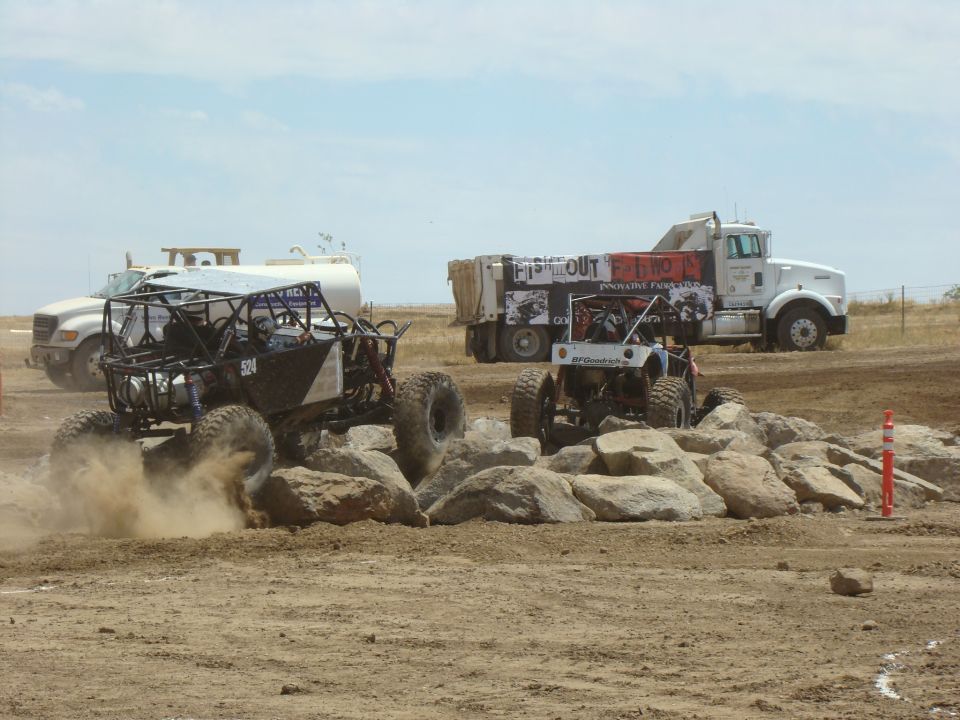 Two custom off-road vehicles navigate a rocky course with a large dump truck in the background