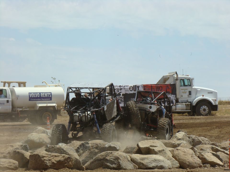 Two off-road buggies race over rocks, with a Volvo Rents truck and a semi-truck in the background
