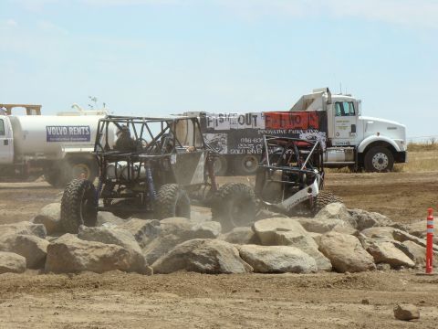 1989 Ford Bronco - Custom off-road vehicles tackle a challenging rock course under a bright sky, with support vehicles nearby.