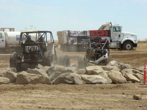 1989 Ford Bronco - Two off-road buggies traverse a rocky obstacle course, with a large dump truck and water truck visible in the background.