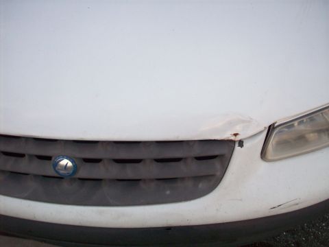 1985 Ford Bronco - Close-up of a white car's front grille and headlight shows damage to the hood.