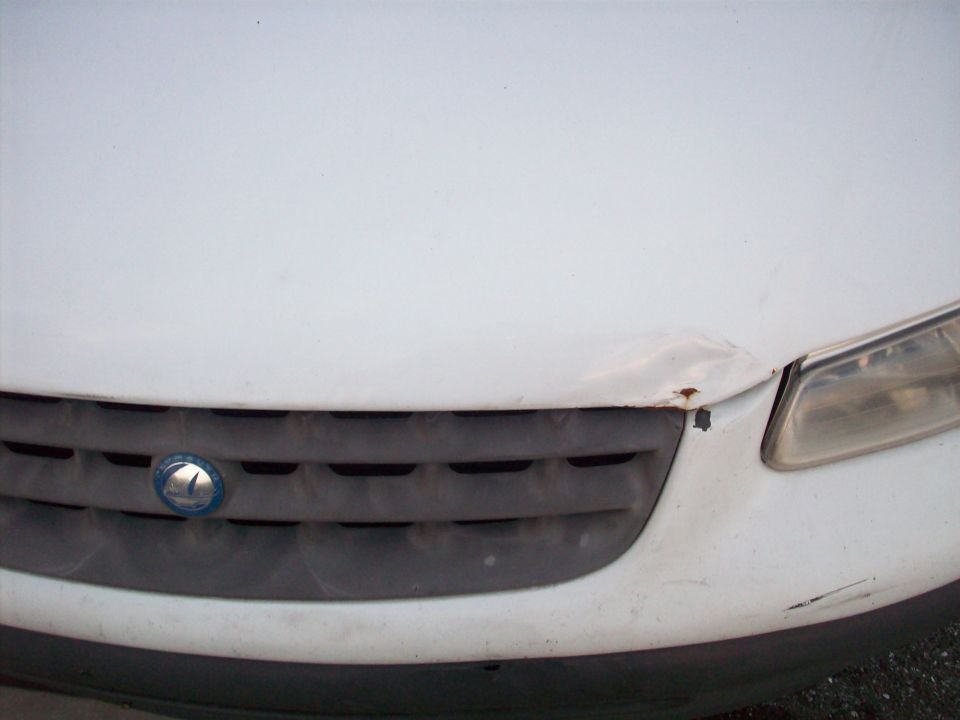 White car front grille with blue emblem and damaged hood with rust near headlight