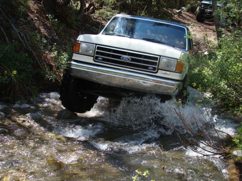 1989 Ford Bronco - A white Ford Bronco navigates a rocky stream in a wooded area, with another vehicle visible in the background.