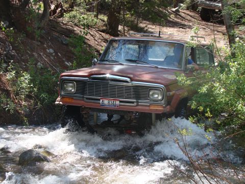 1989 Ford Bronco - Jeep Grand Wagoneer navigates a rocky stream bed in a wooded area.
