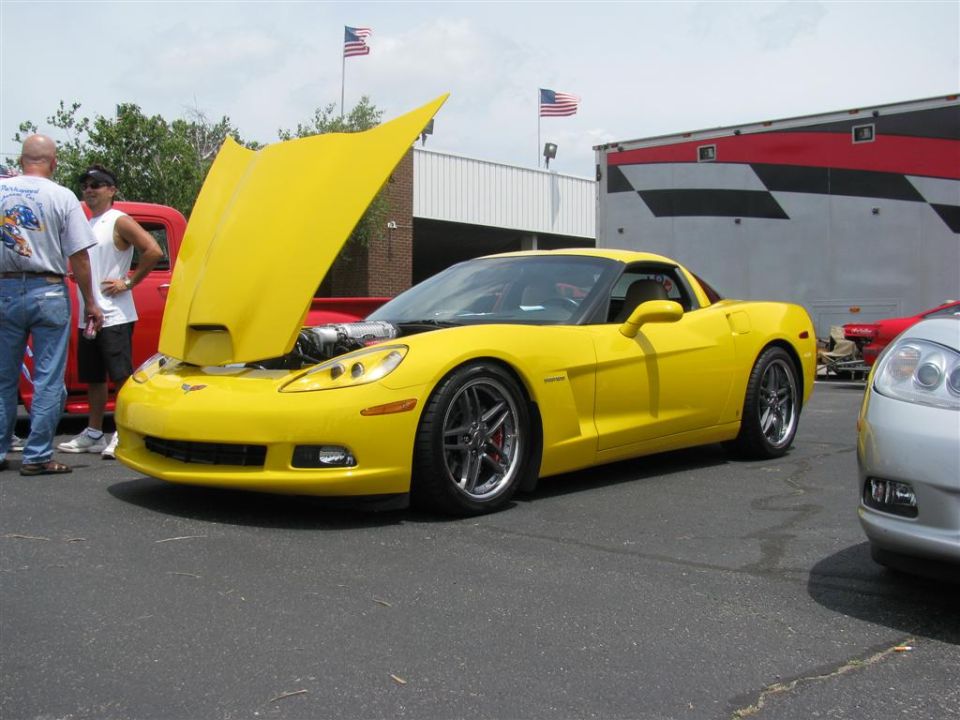 Yellow Chevrolet Corvette with hood open, showing engine. Two men stand nearby. American flags fly in background