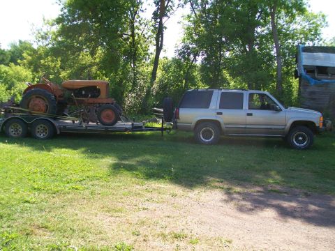 2000 Chevrolet Tahoe - An SUV tows a tractor on a trailer, with a wooden structure in the background.