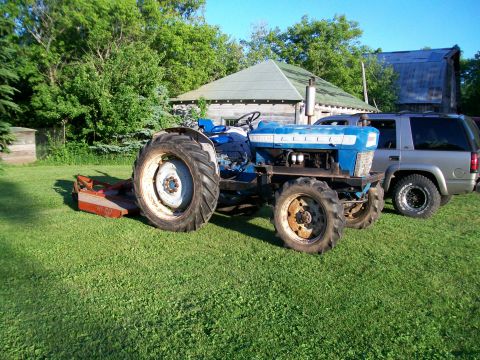 2000 Chevrolet Tahoe - A blue tractor with a mower attachment is parked on a grassy lawn next to a silver SUV.