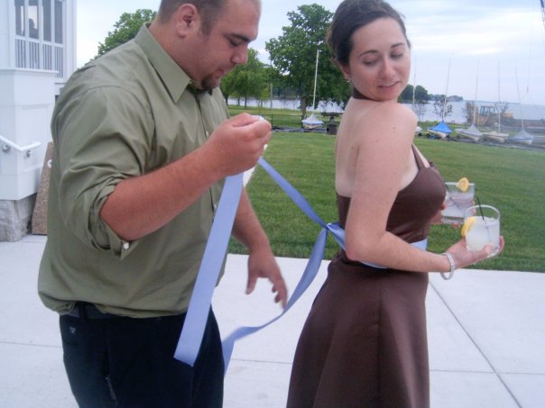 Man holding blue ribbon and woman in brown dress with a drink outdoors near water
