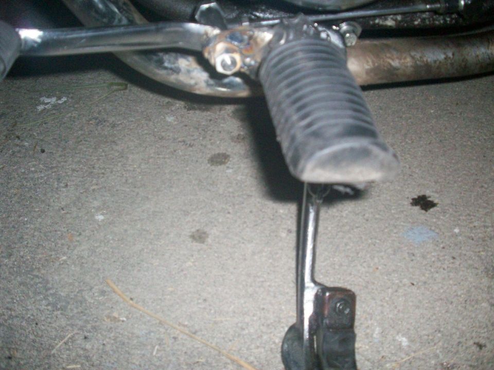 Close-up of a motorcycle rubber footpeg on a chrome mount with weld marks, lever extending below, over concrete.