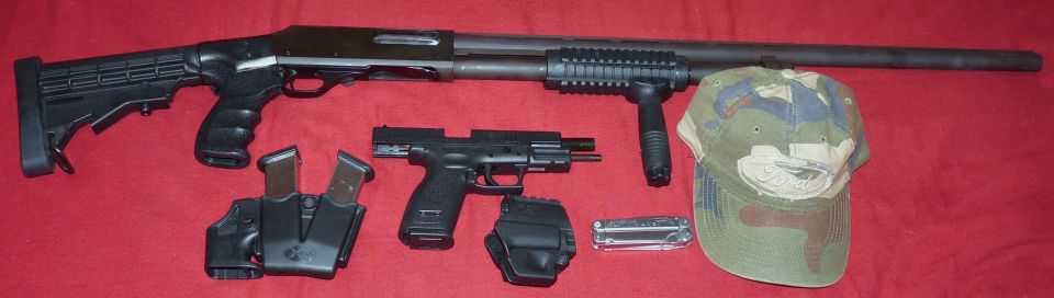 Shotgun, pistol, magazines, holsters, multi-tool, and camouflage hat laid out on a red surface