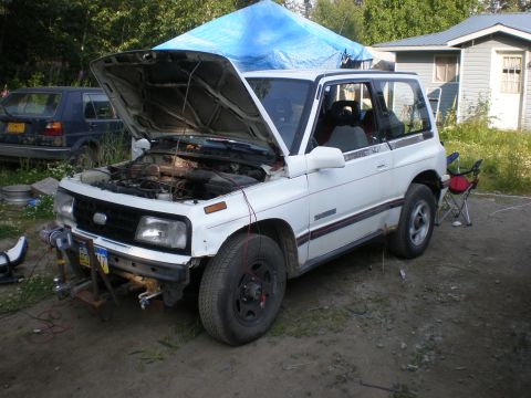 1987 Suzuki Samurai - A white 1987 Suzuki Samurai is parked outdoors with its hood open, partially covered by a blue tarp.