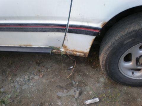 1987 Suzuki Samurai - Close-up of a white vehicle's side door and wheel, revealing extensive rust damage on the lower body panel.