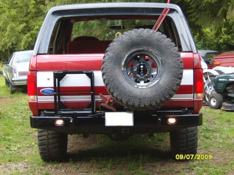 1989 Ford Bronco - The rear of a red Ford Bronco is shown, a spare tire and a tow hitch. A second vehicle is visible to the left.