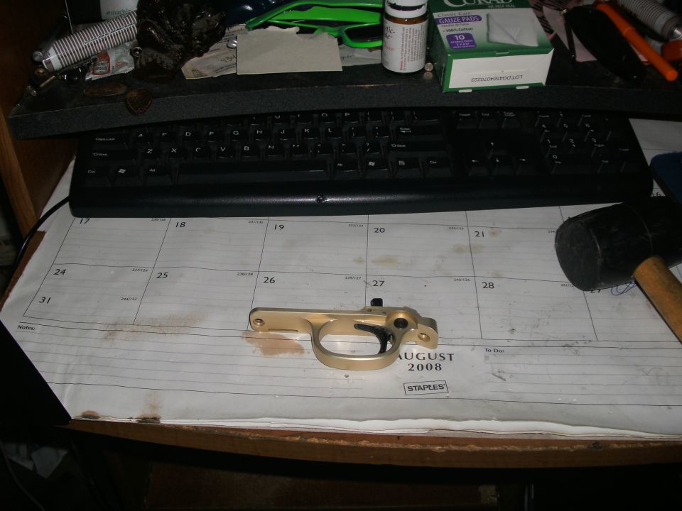 Gold rifle trigger guard assembly on an August 2008 desk calendar beside a keyboard and rubber mallet