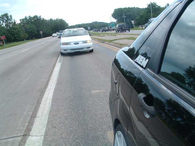 black car's side with an AEM sticker on the rear window, driving on a road next to a white car