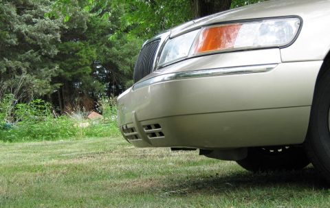 2000 Mercury Grand Marquis - The front of a gold 2000 Mercury Grand Marquis is visible, parked on grass with trees in the background.