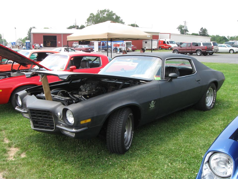 Black 1970s muscle car with hood open, showing engine. Another red car is visible behind it