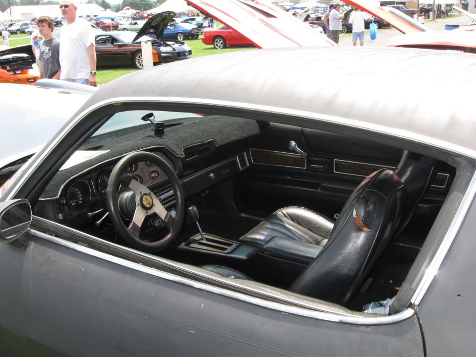 Interior of a dark car with bucket seats, steering wheel, and dashboard visible. Other cars and people are in the background