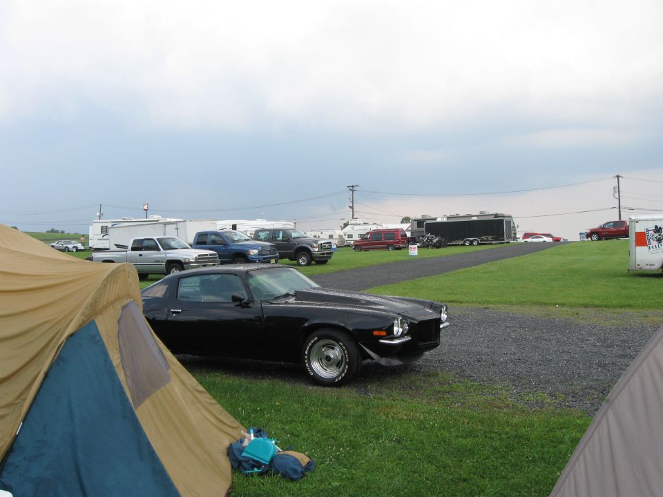 Black coupe with a prominent hood scoop and aftermarket wheels parked on gravel, viewed from the front three-quarter angle