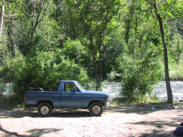 Side profile of blue lifted Ford pickup parked by a river with dense green trees