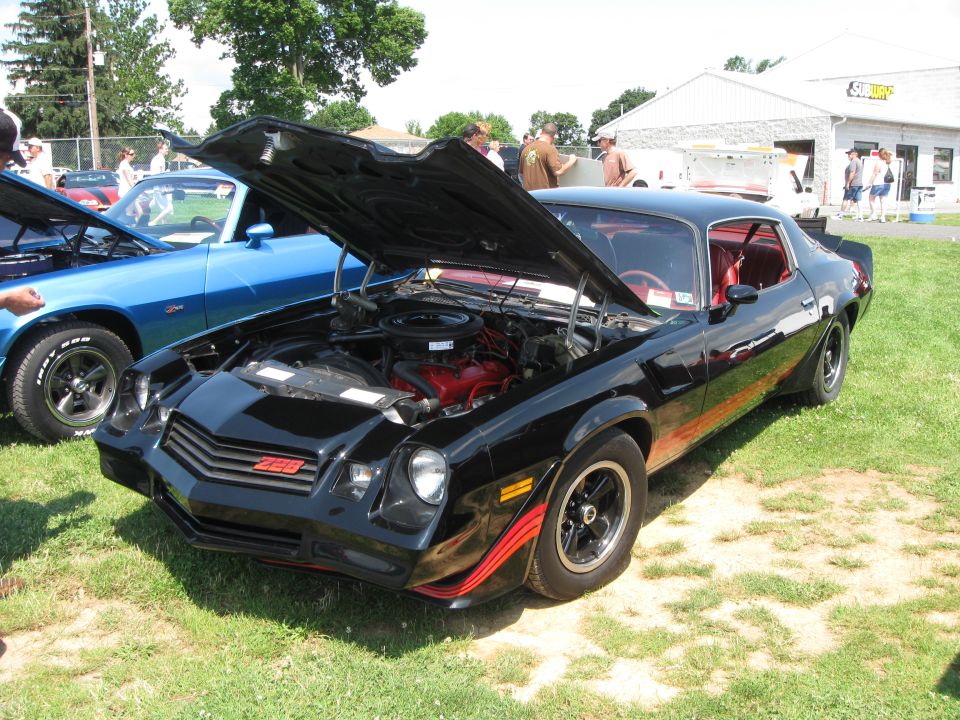 black Chevrolet Camaro Z28 with hood open, parked on grass next to a blue car