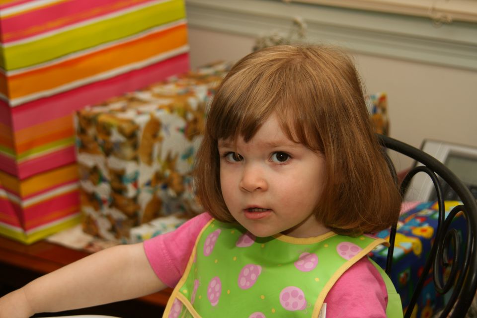 Child wearing a pink shirt and green bib with pink circles sits in a chair, with gift-wrapped boxes in the background