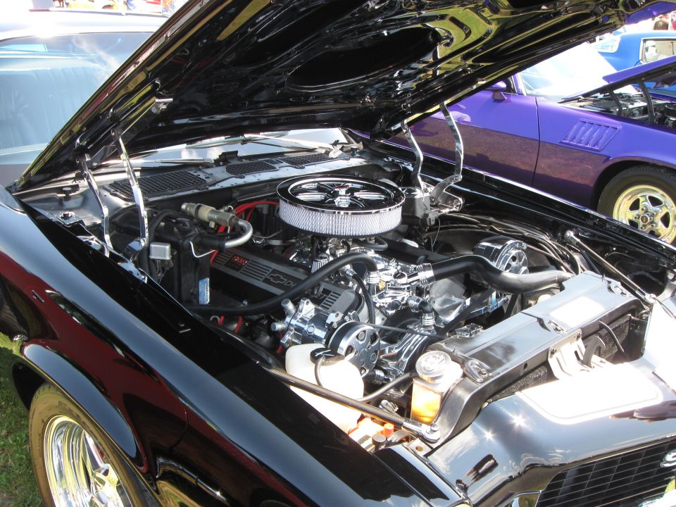 Engine bay of a black classic car with chrome air cleaner and Chevrolet valve covers. Purple car visible in background