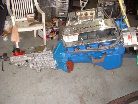 1970 Ford Torino - A Ford Racing engine and transmission assembly is shown on a concrete floor, with various parts and a chair in the background.