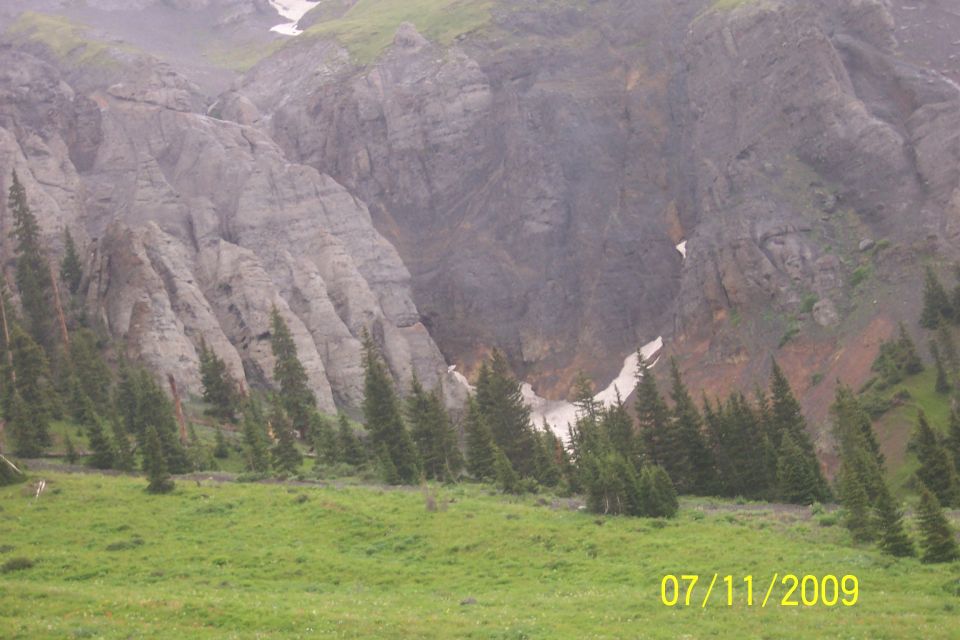 Mountain landscape with rocky cliffs, green grass, and scattered pine trees. A small patch of snow is visible on a cliff face