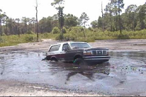 1993 Ford Bronco - A 1993 Ford Bronco navigates through a muddy, water-filled area with trees in the background.