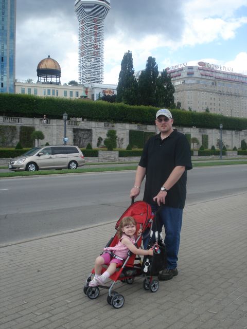 2000 Ford Crown Victoria - A gold minivan is visible on the left side of the frame, parked near a sidewalk where a man is pushing a child in a stroller.