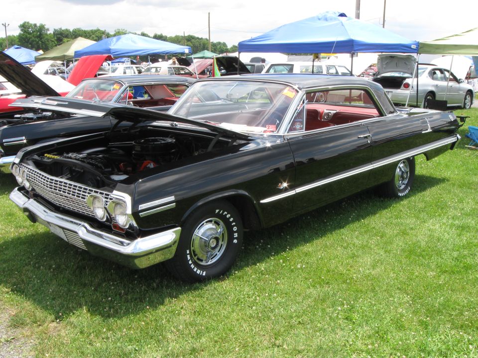 Black 1963 Chevrolet Impala coupe with red interior, hood open, parked on grass at an outdoor car show