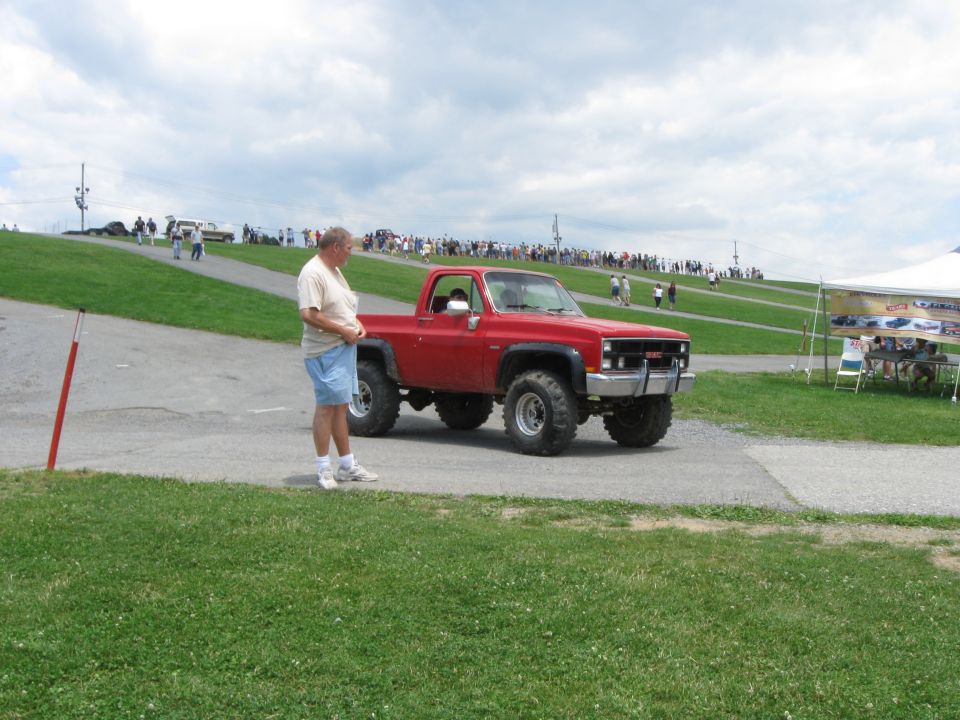 Red GMC pickup truck with lifted suspension and large tires, parked on a paved surface with a man standing nearby. A crowd and tents are visible in the background