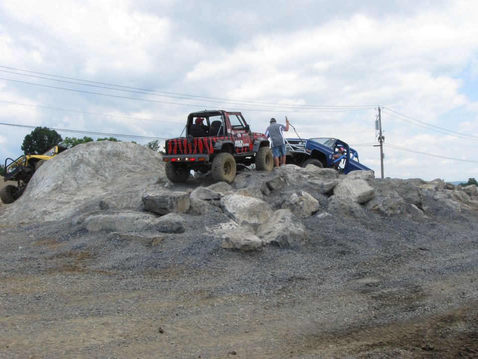 Red Jeep and blue vehicle on rocks, yellow vehicle on left. Person with rope near red Jeep