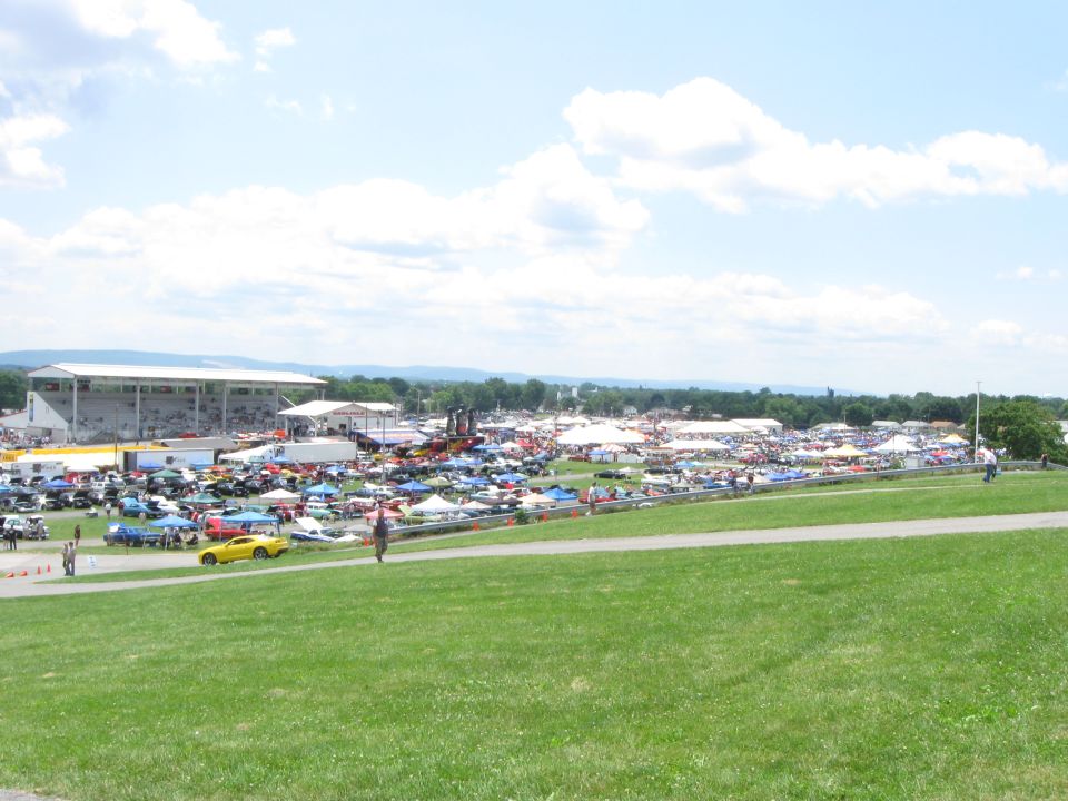 Wide view of a car show with numerous cars parked under tents and in open areas, a grandstand visible in the background
