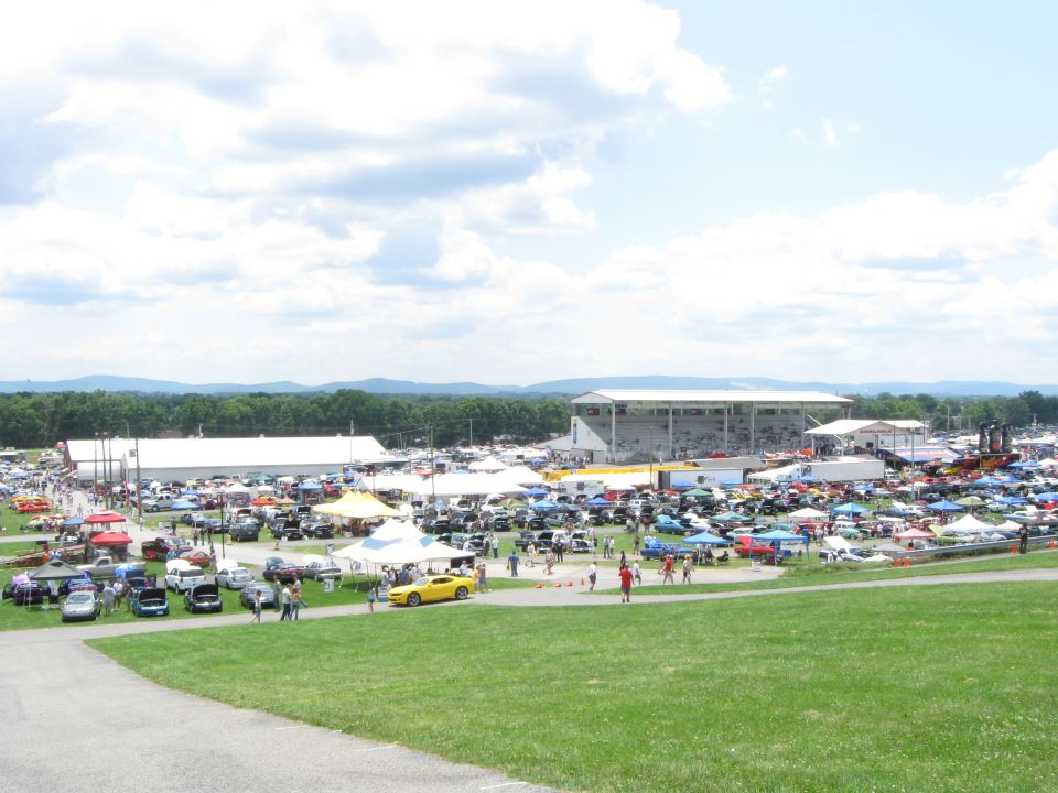 Outdoor car show with hundreds of vehicles parked under tents and in open lots, stadium in background
