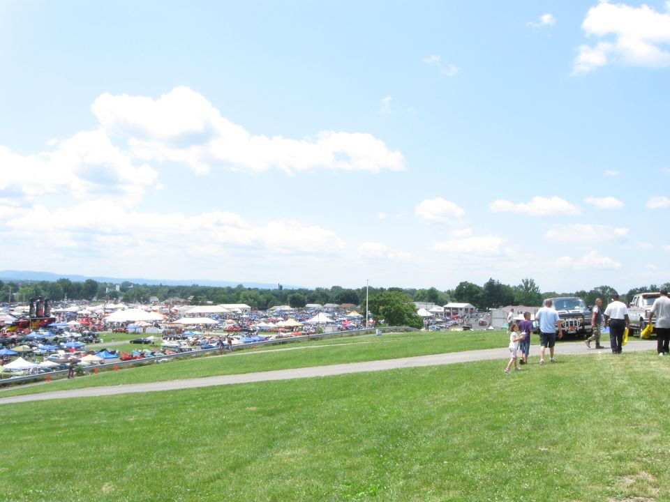 Wide shot of a car show with numerous tents and vehicles spread across a grassy field under a partly cloudy sky