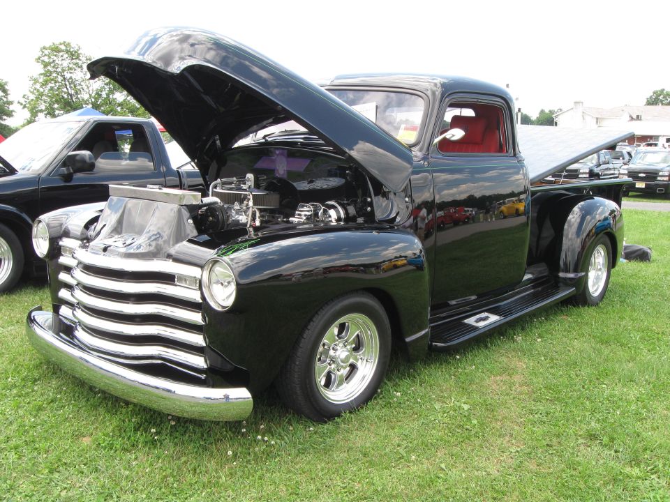 Black 1950s pickup truck with chrome grille and hood open, showing engine. Another black pickup is parked to the left