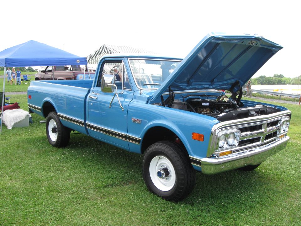 Blue pickup truck, hood open, 3q-front view. Another truck visible under a tent