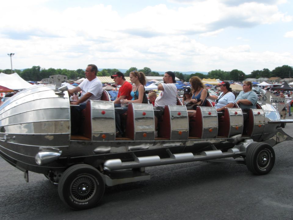 Custom-built amusement ride vehicle with multiple rows of seats, carrying several passengers. It has a silver, segmented body and is on display outdoors