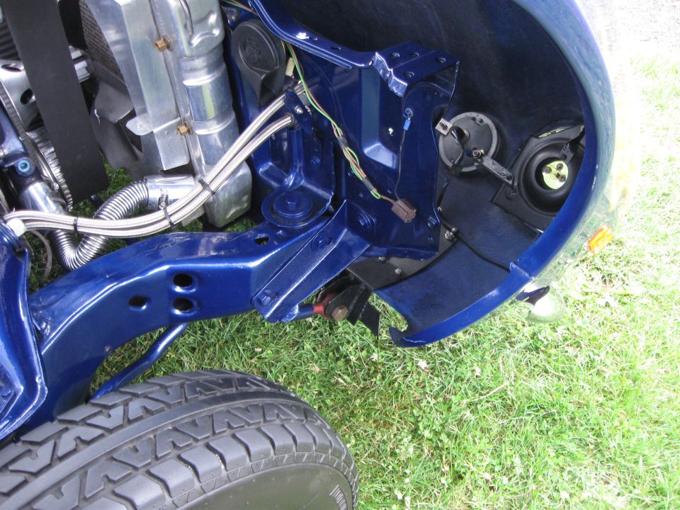Close-up of a blue car's front chassis, showing the radiator, engine mounts, and wiring. A tire is visible in the foreground