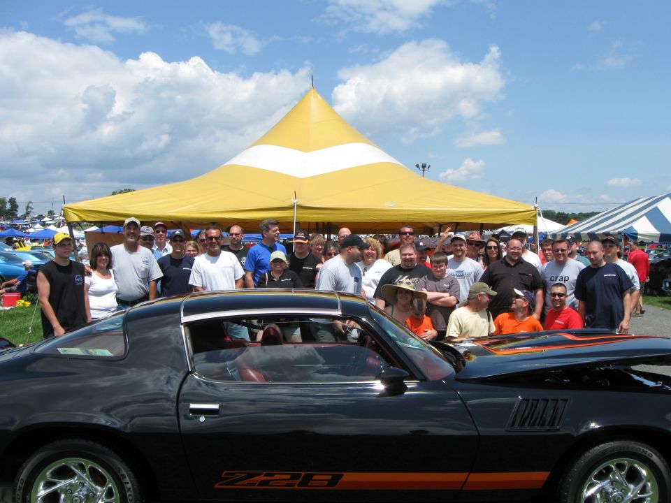 Black Z28 Camaro with orange stripes, hood open, parked in front of a crowd under a yellow tent