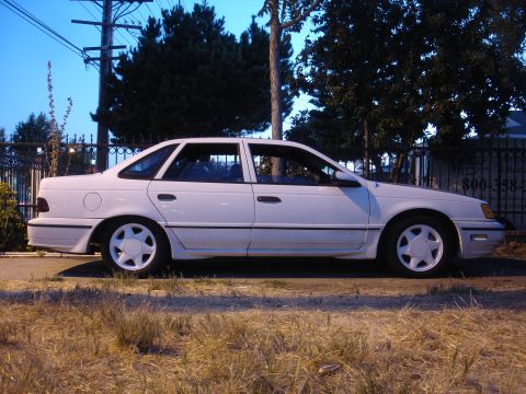1990 Ford Taurus - This white 1990 Ford Taurus is parked on the side of the road with trees and a fence in the background.