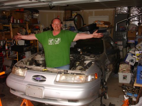 1992 Ford Taurus - A man stands with his arms outstretched in the engine bay of a silver Ford Taurus, surrounded by garage clutter.