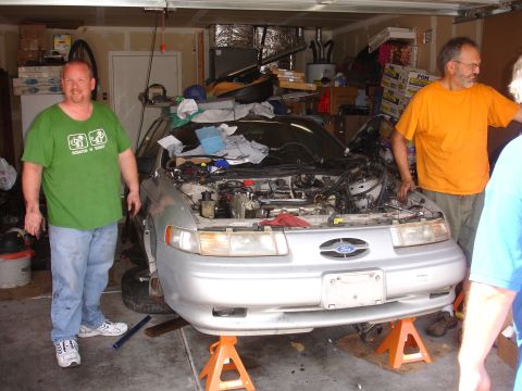 1992 Ford Taurus - Two men are working on a silver car that is raised on jack stands in a garage, with the engine compartment open.