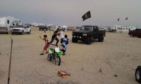 1995 Ford Bronco - A black 1995 Ford Bronco with a pirate flag flying overhead is parked on the sand, with people and a dirt bike nearby.